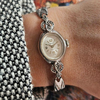 Silver watch with decorative charm on a wrist against a patterned fabric background